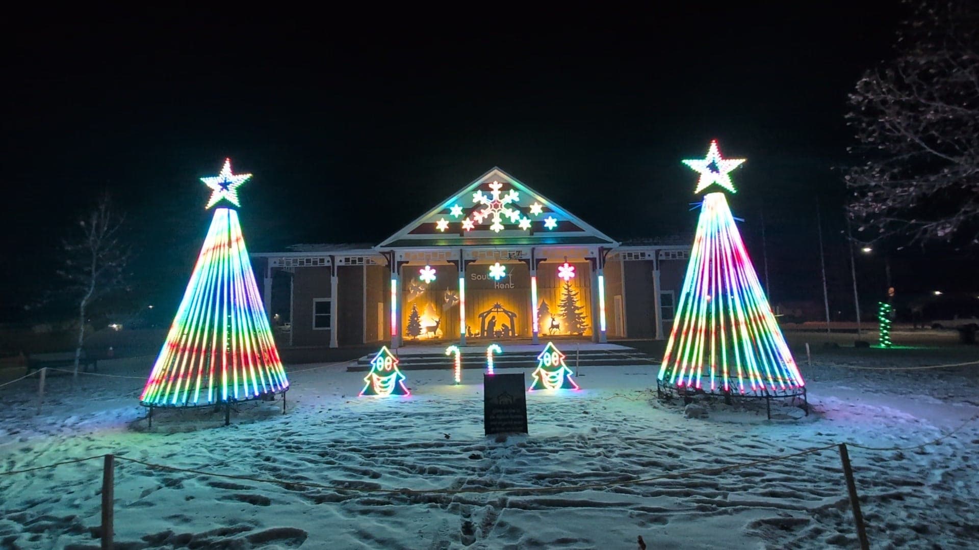 Rainbow-striped pixel mega trees beside the lit pavilion stage on a snowy lawn